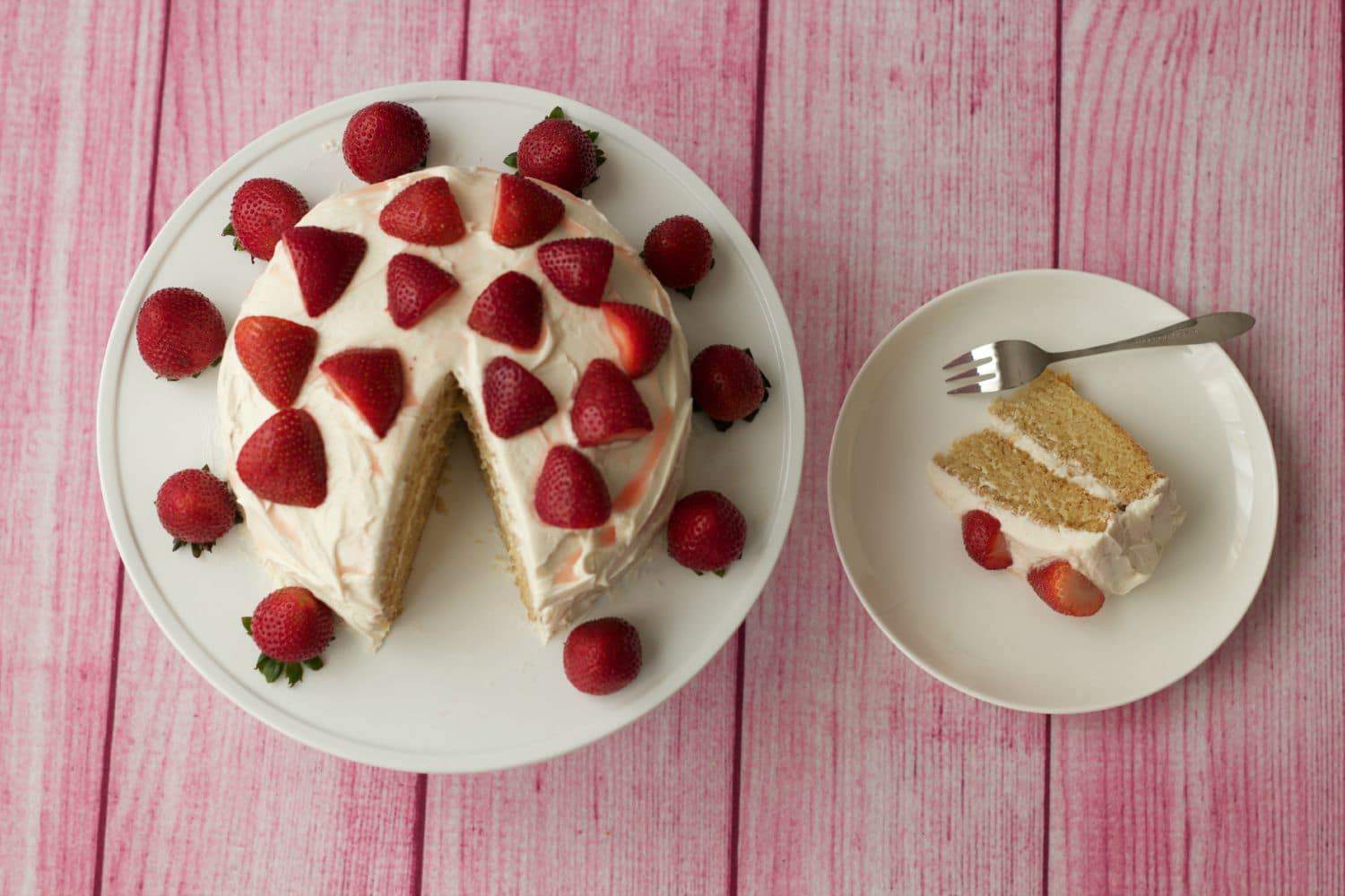Vegan vanilla cake topped with fresh strawberries and a slice of vanilla cake on a white plate with a cake fork. 