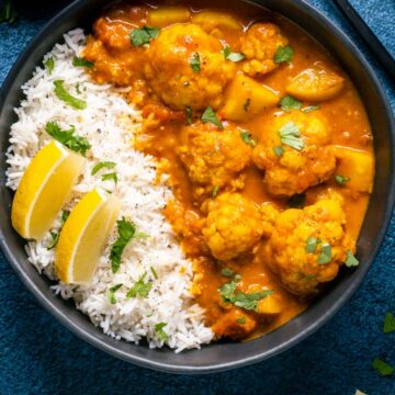 Vegan cauliflower curry with rice and lemon wedges in a black bowl.