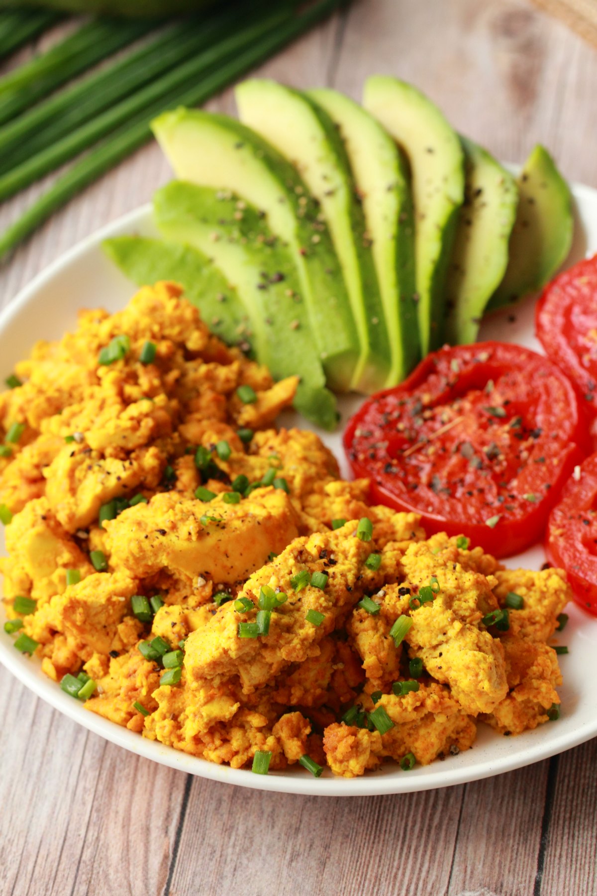 Tofu scramble topped with chopped chives on a white plate with fried tomato and sliced avocado. 